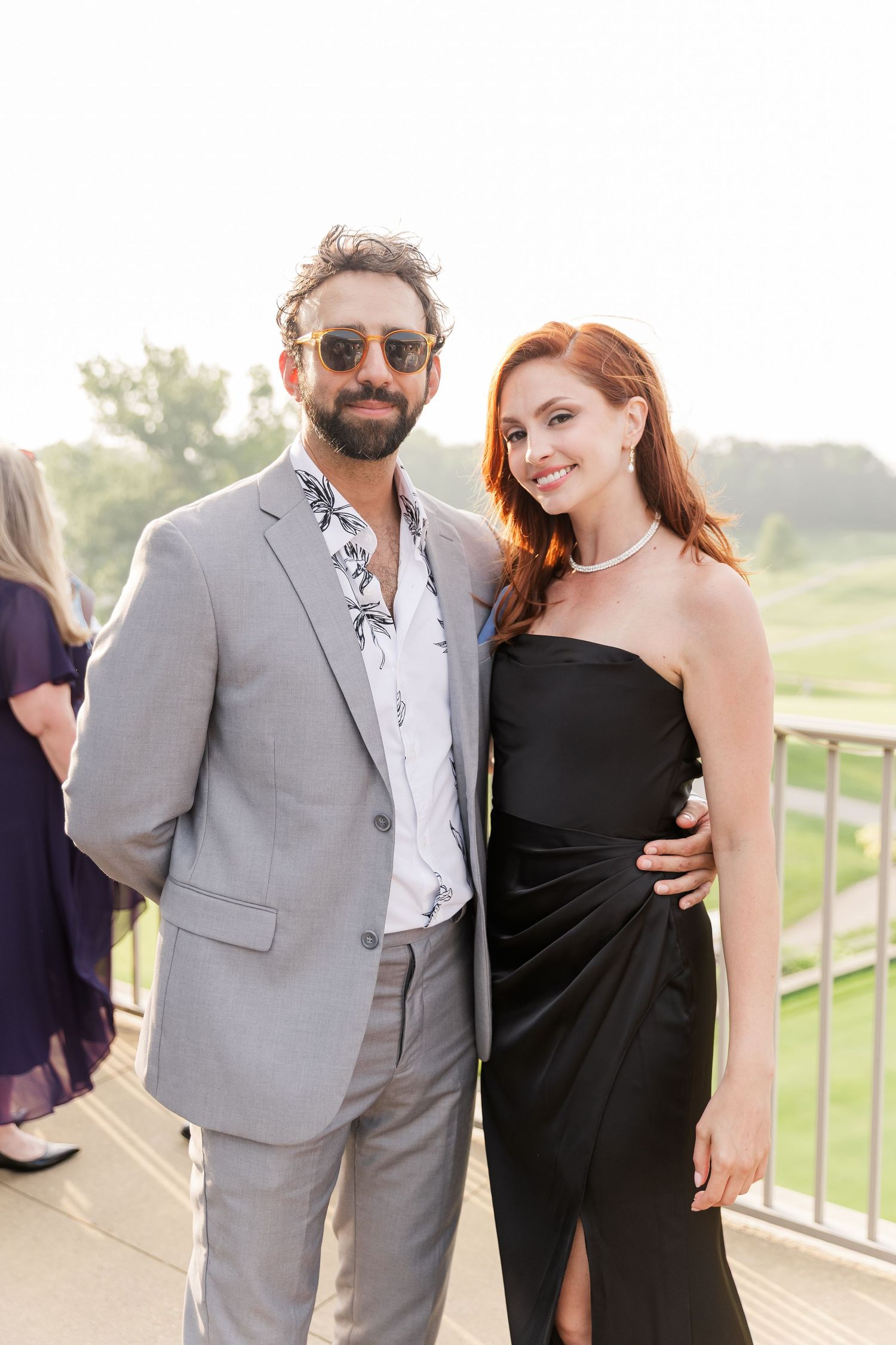 Smiling couple posing together outdoors at wedding reception, man in gray suit, woman in black dress.