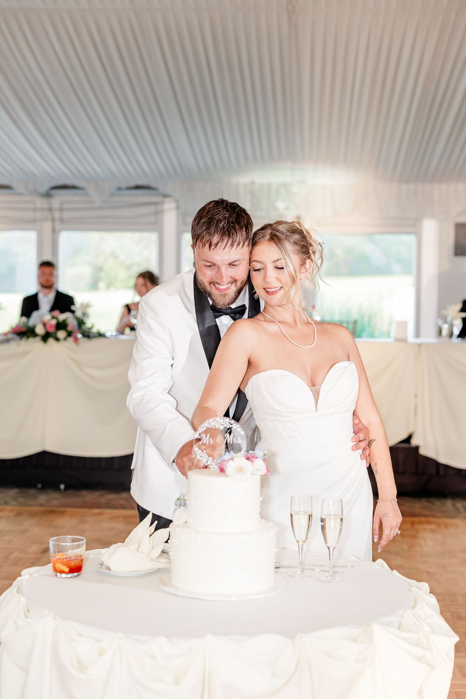 Bride and groom cutting white two-tier wedding cake at reception with champagne glasses nearby.