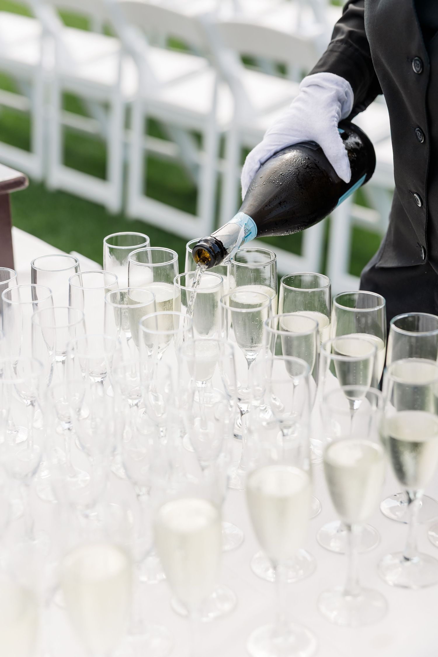 A white-gloved waiter pours champagne into elegant flutes arranged in rows at an outdoor wedding reception.