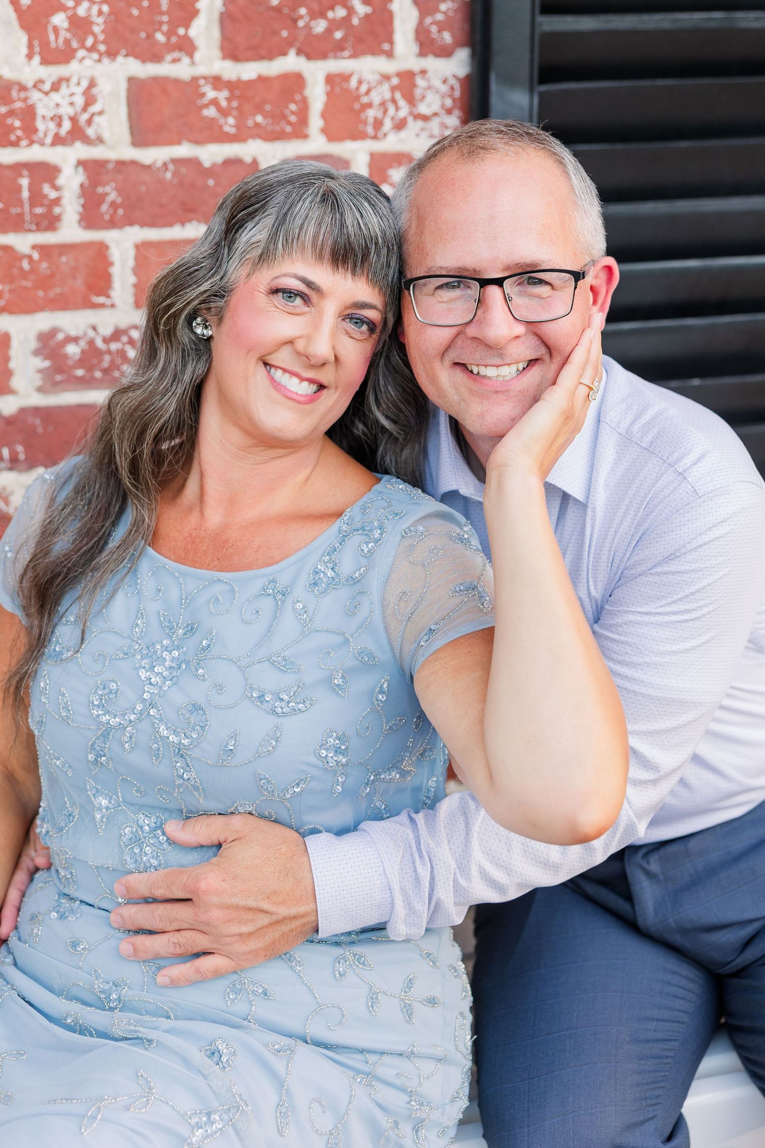 Smiling middle-aged couple posing together against a brick wall, woman in blue lace dress and man in glasses.