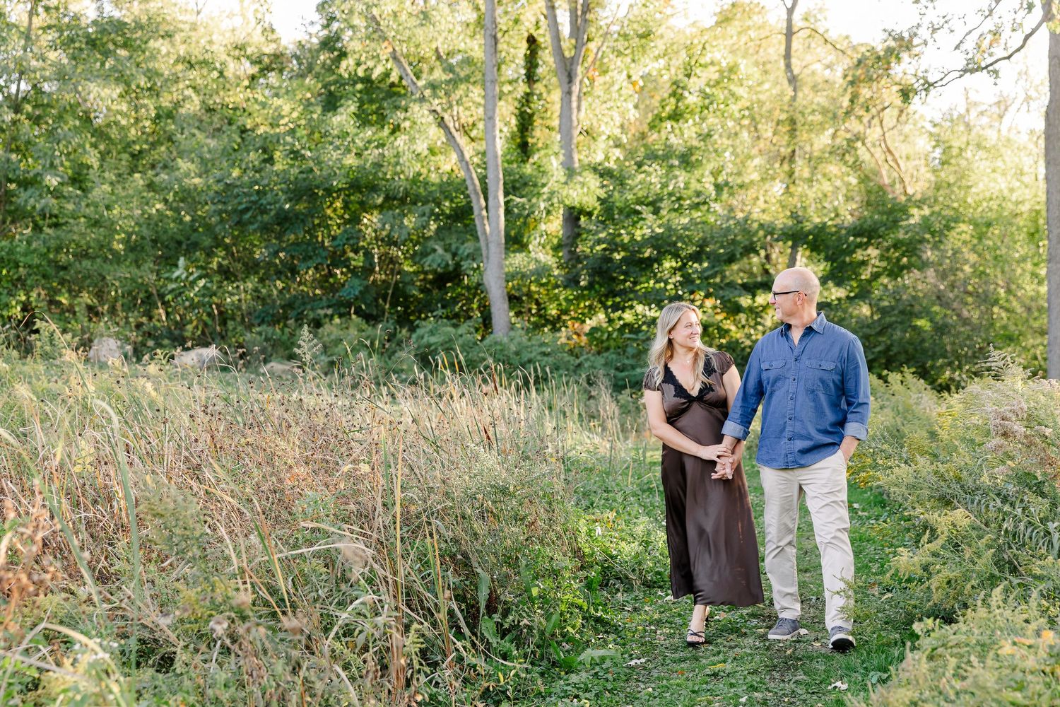 Couple holding hands among backyard trees and tall grasses during fall portraits in St. Charles, IL.