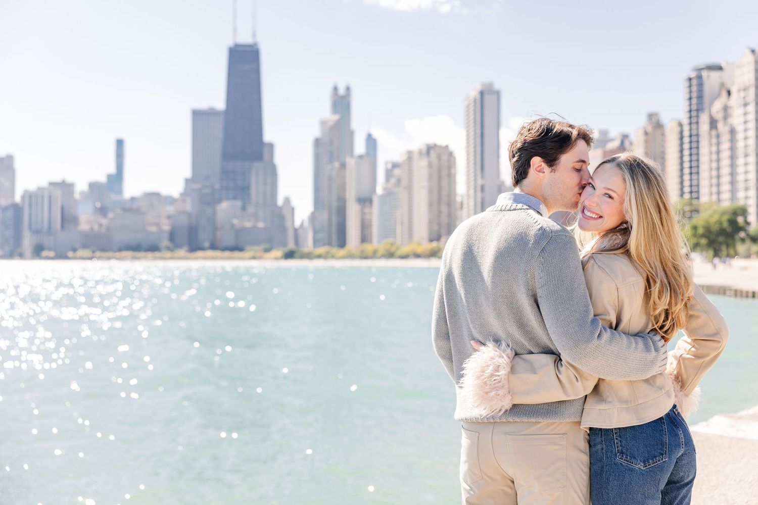 Couple embracing and smiling on Chicago lakefront with city skyline and John Hancock Center in background.