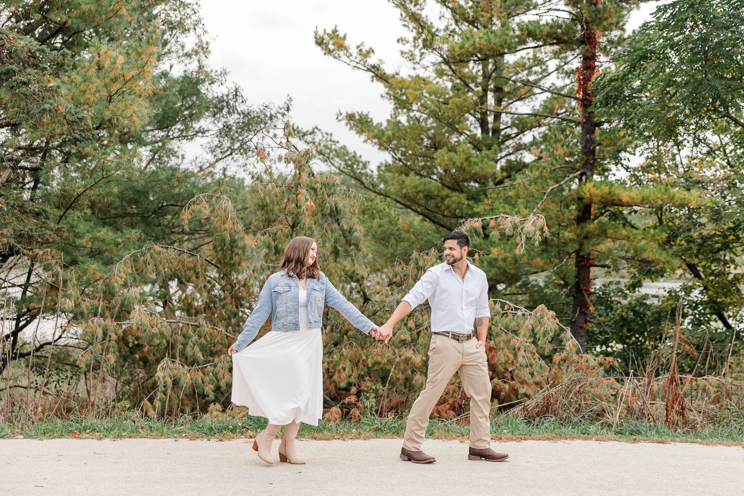 Smiling couple holding hands and walking along a gravel path surrounded by pine trees during an outdoor engagement photo session.