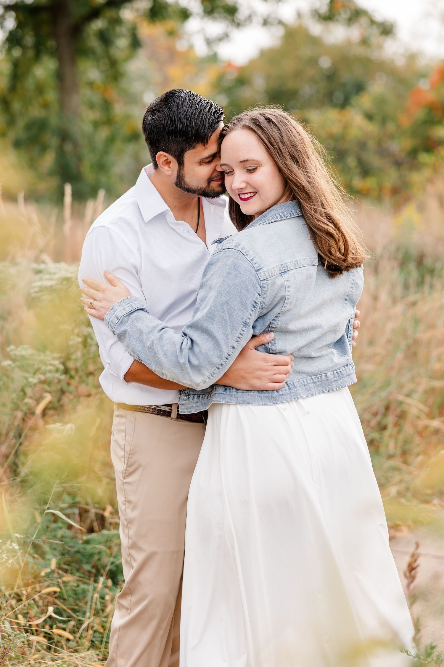 Man in white shirt kisses smiling woman in denim jacket and white skirt as they embrace in a golden autumn meadow.