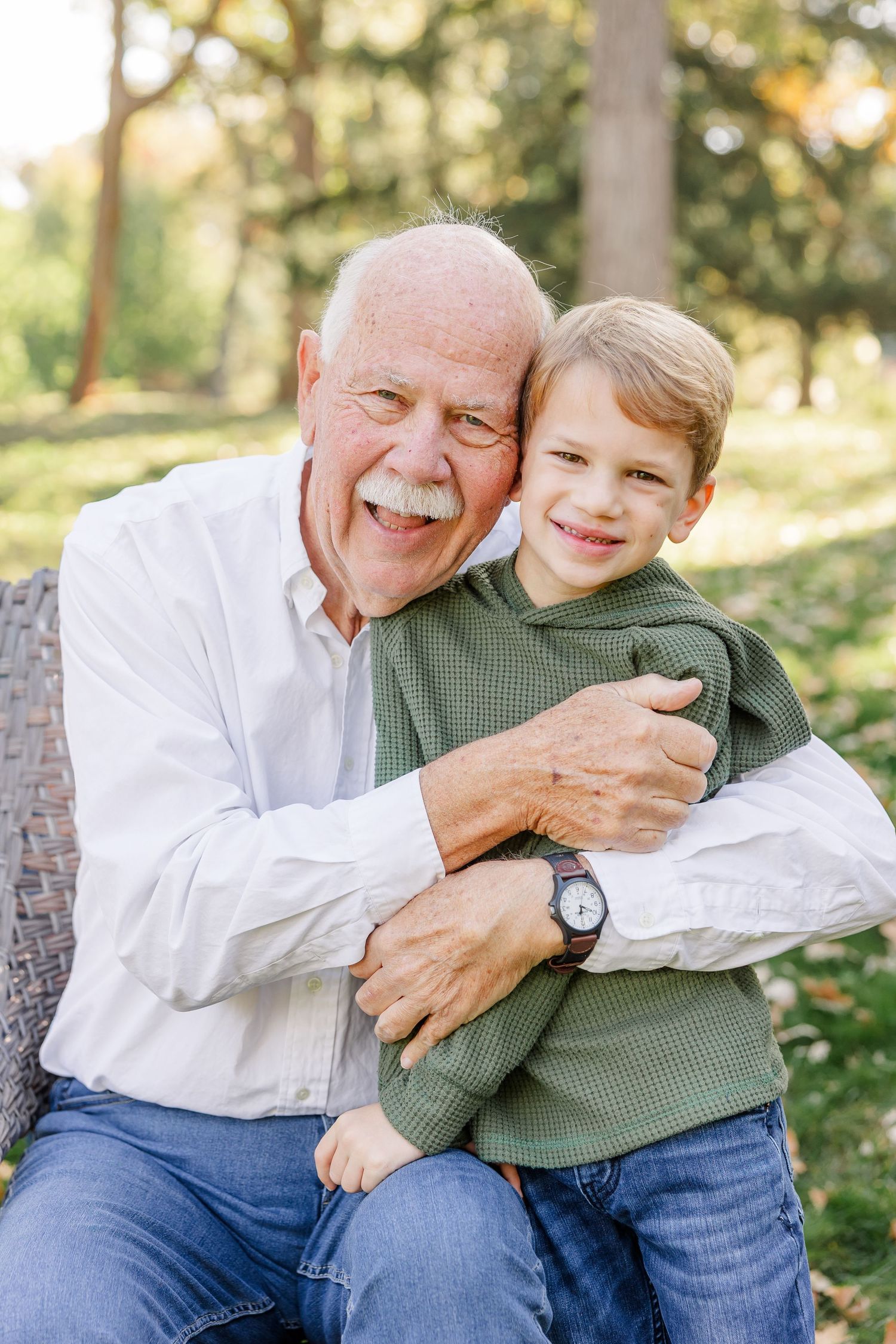 Elderly grandfather hugging young grandson outdoors, both smiling joyfully in a lush green park setting.