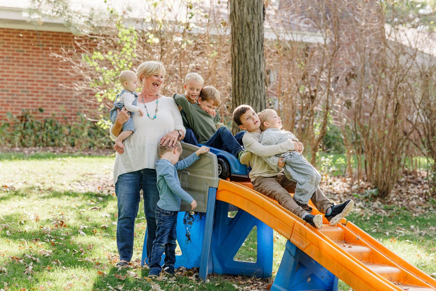A grandmother plays with five grandchildren on a colorful backyard slide during a fall family gathering.