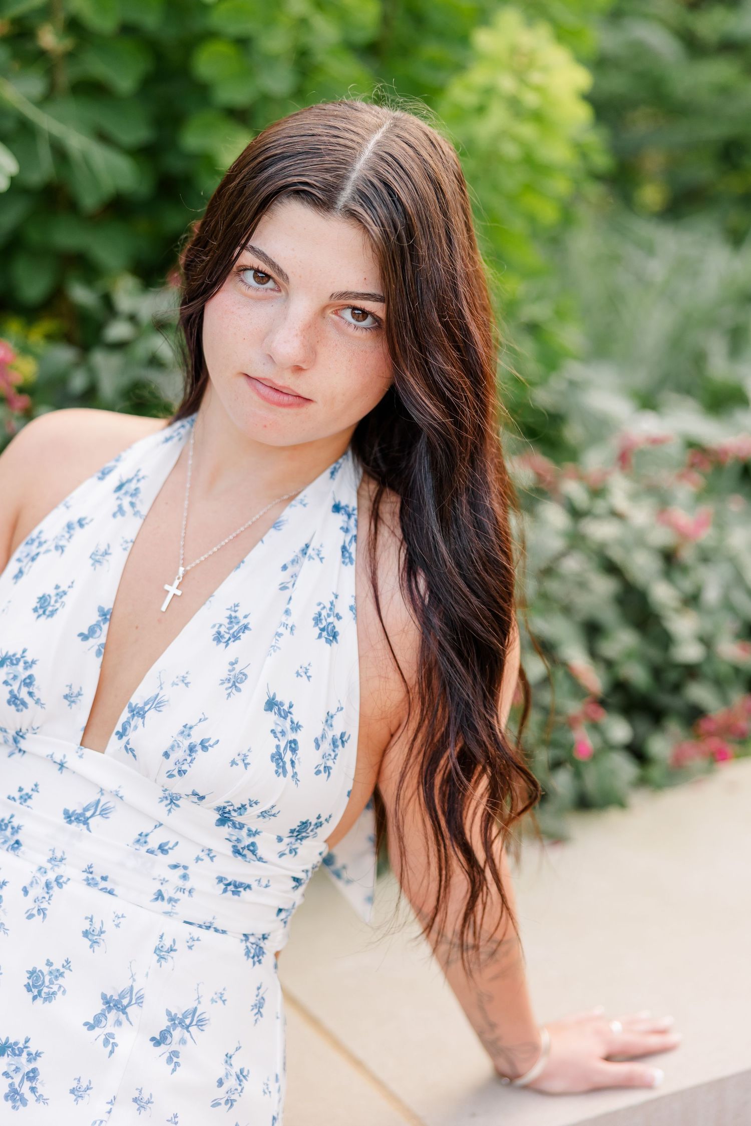 Young brunette woman in white blue floral halter dress posing outdoors wearing cross necklace in garden setting.