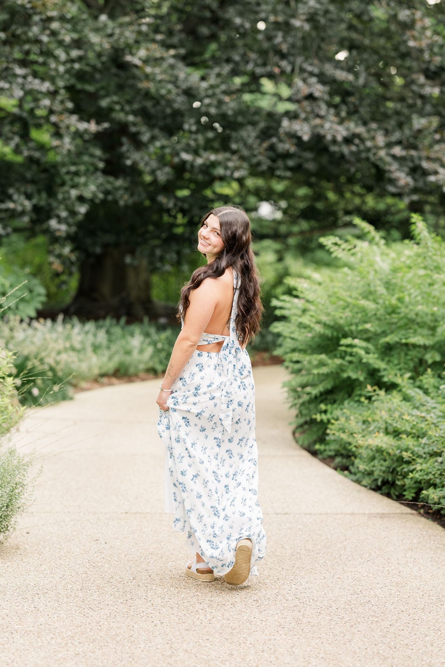 Woman in floral maxi dress walking garden path, smiling over shoulder, lush greenery background.