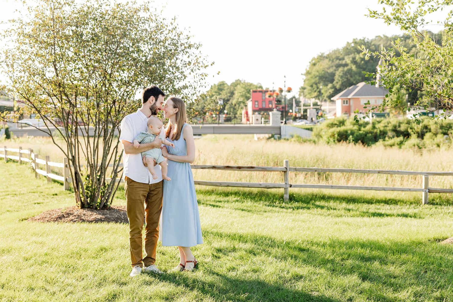 A couple shares a tender moment with their baby in a sunlit park near a white fence and lush greenery.