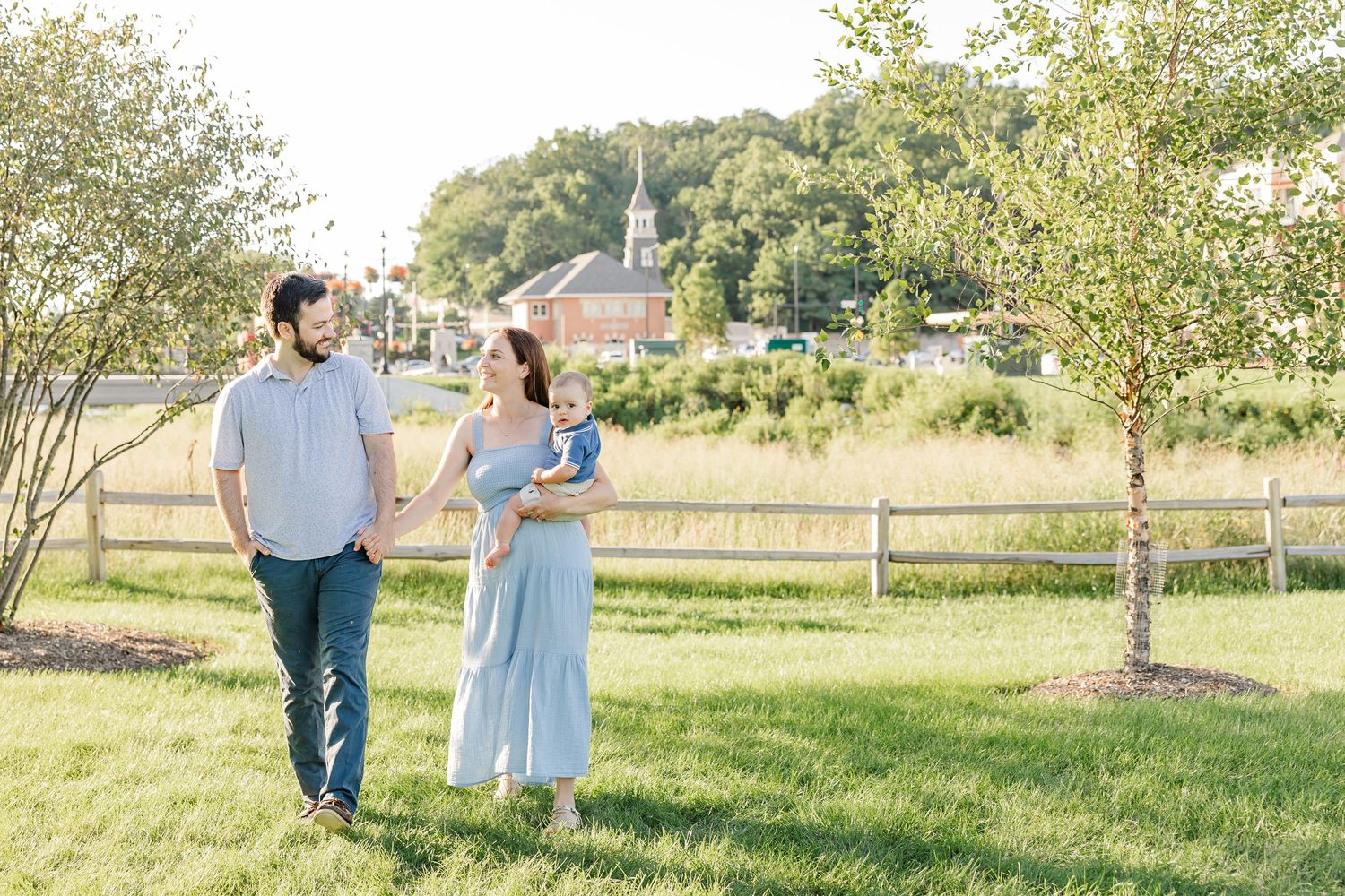 A family strolls through a sunny green park, mother in blue dress holding baby, father beside her, with a white church in background.