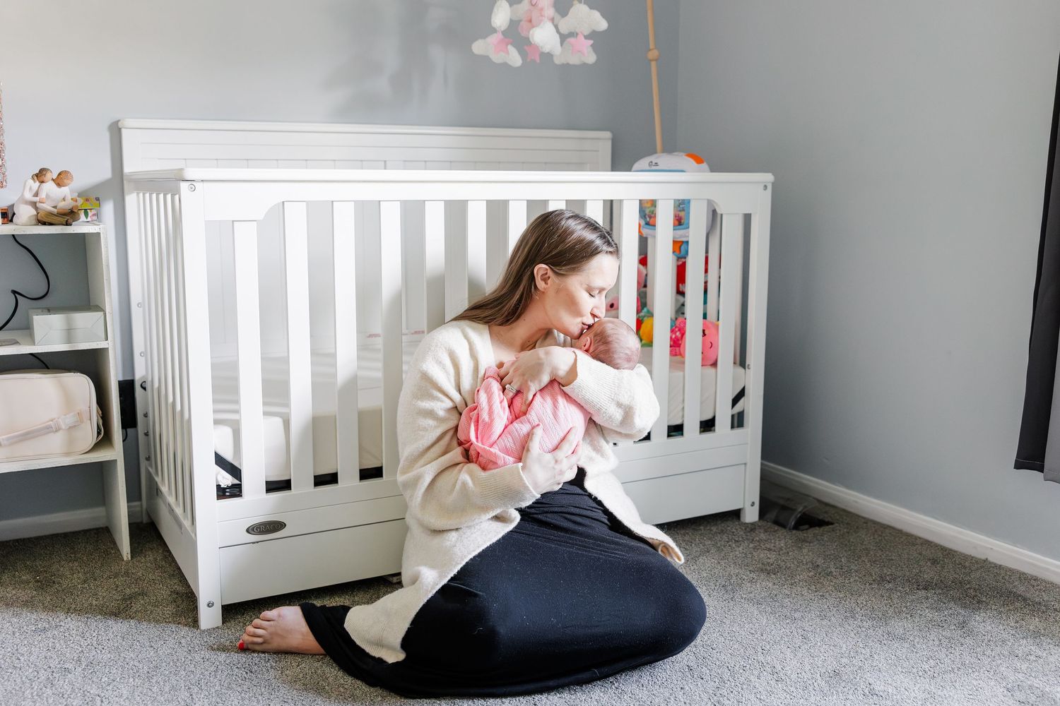 A mother sits on the nursery floor cradling her newborn baby in front of a white crib.
