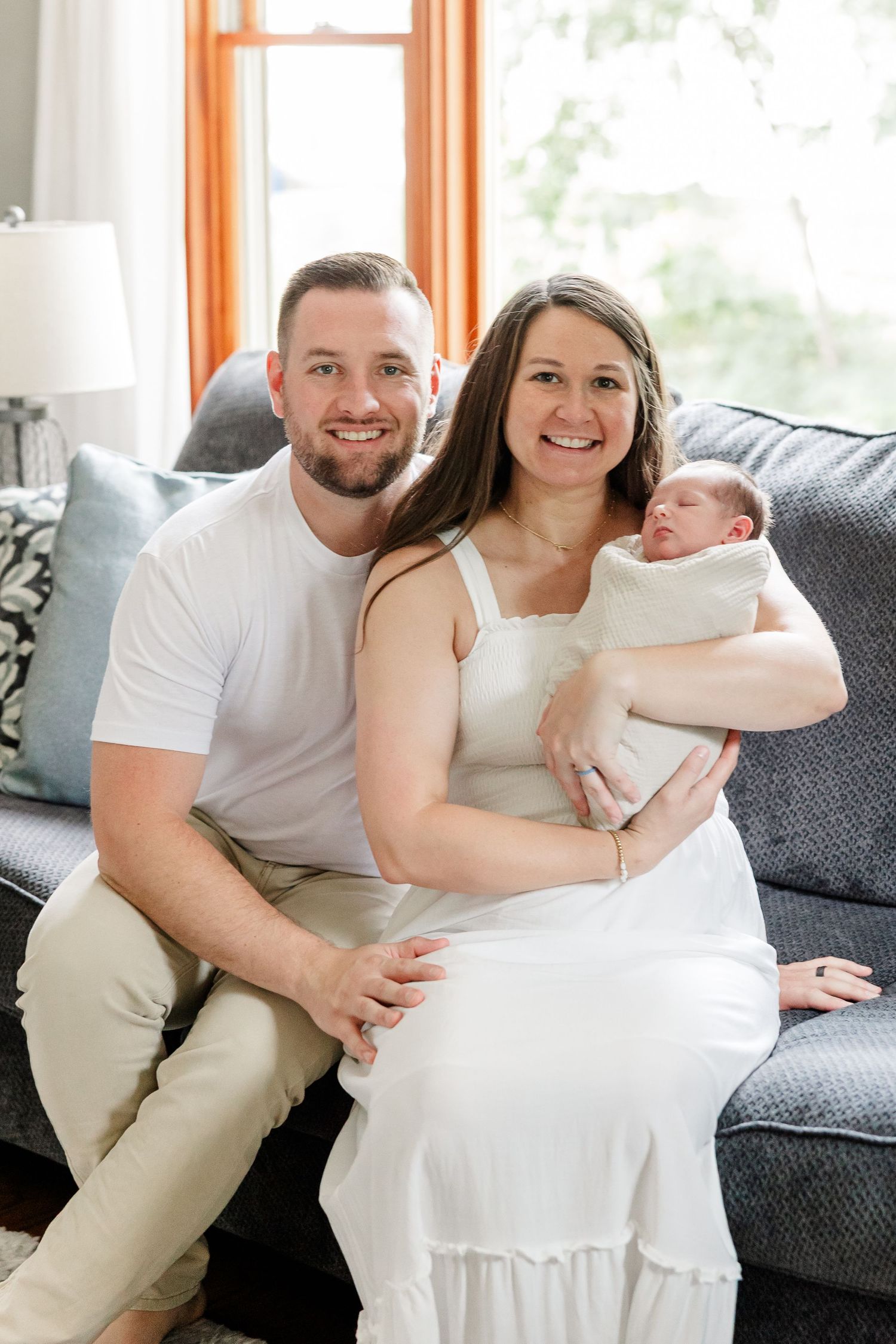 A smiling couple sits on a gray sofa, the woman in white holding a swaddled newborn baby at an in-home newborn photo session.
