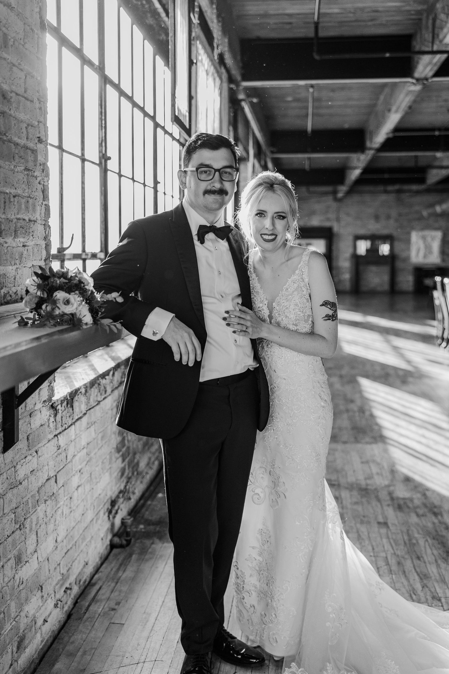 A couple in formal wedding attire poses in a sunlit industrial loft space with brick walls.