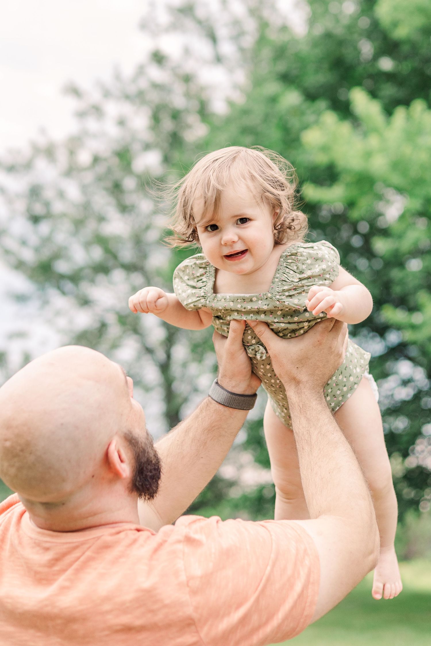 A joyful toddler in a green outfit being lifted up by someone in a peach-colored shirt outdoors.