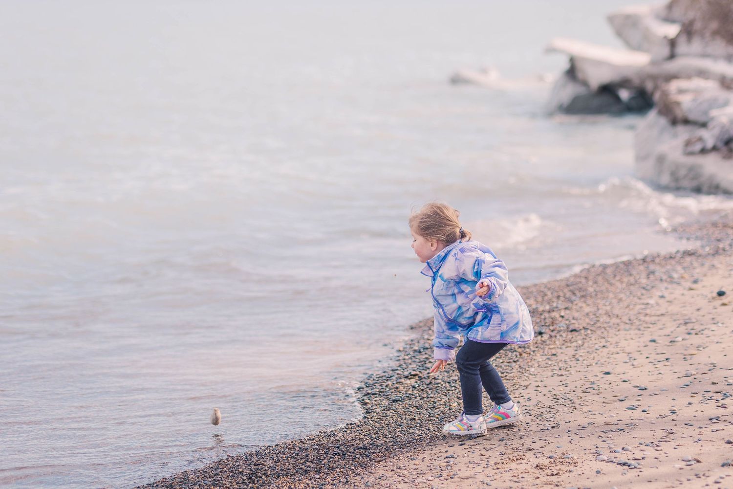 Child in light blue jacket bends down to explore pebbly beach near water.