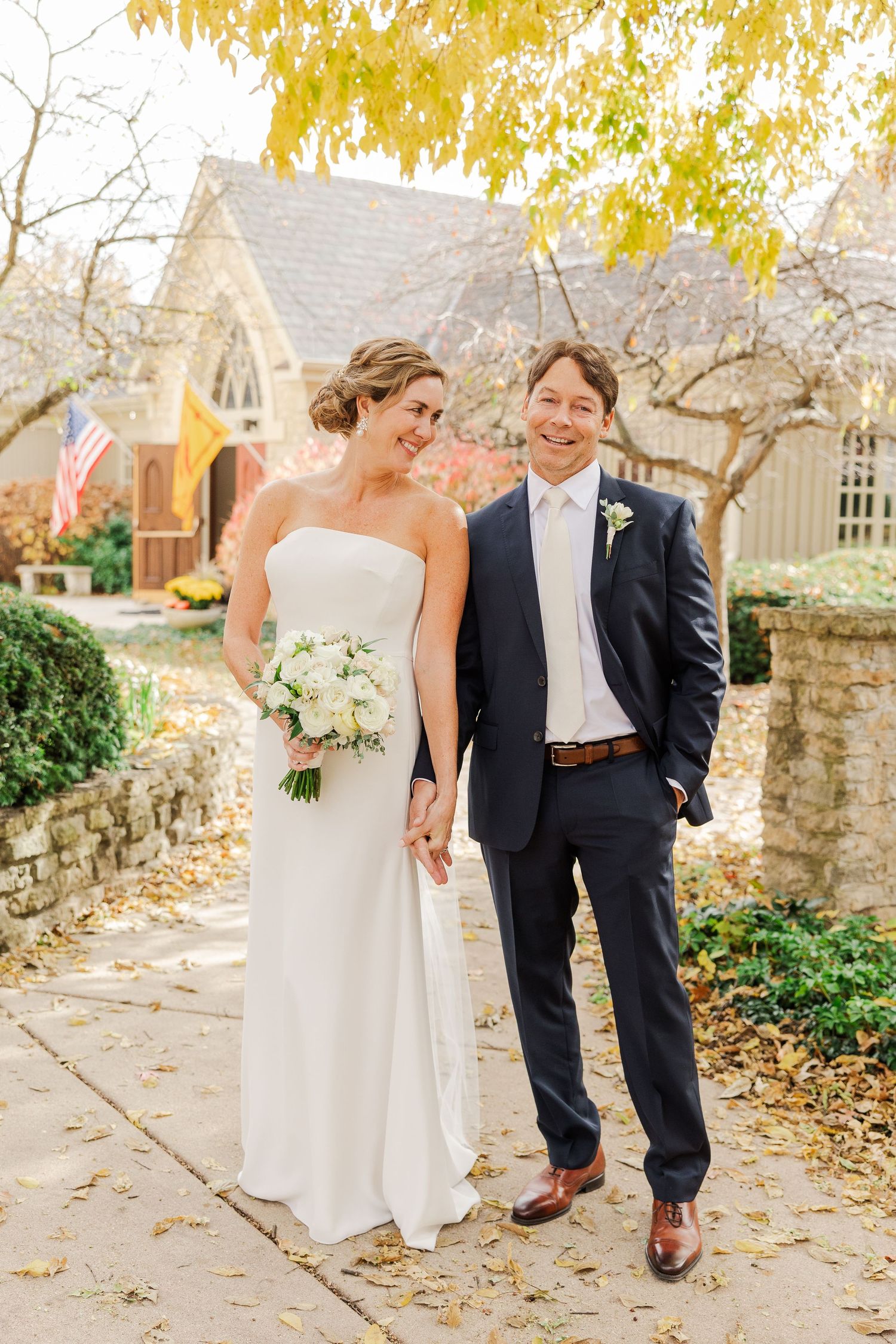 A joyful wedding portrait in front of a stone church with autumn leaves overhead.