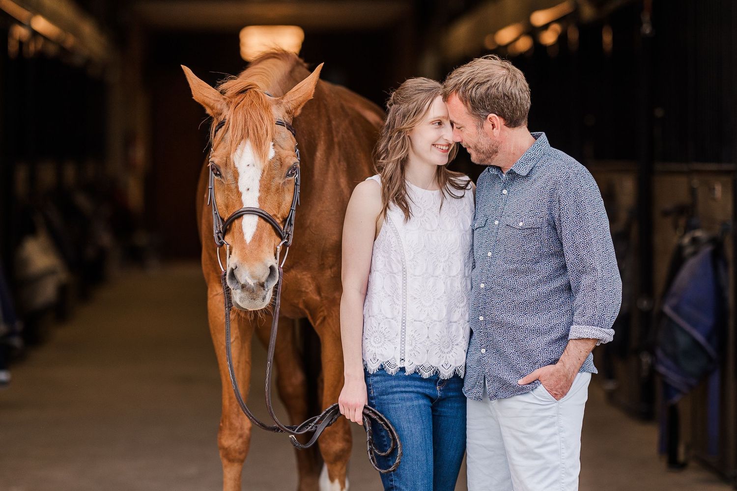 A couple stands close together in a rustic barn stable with a chestnut horse in the background.
