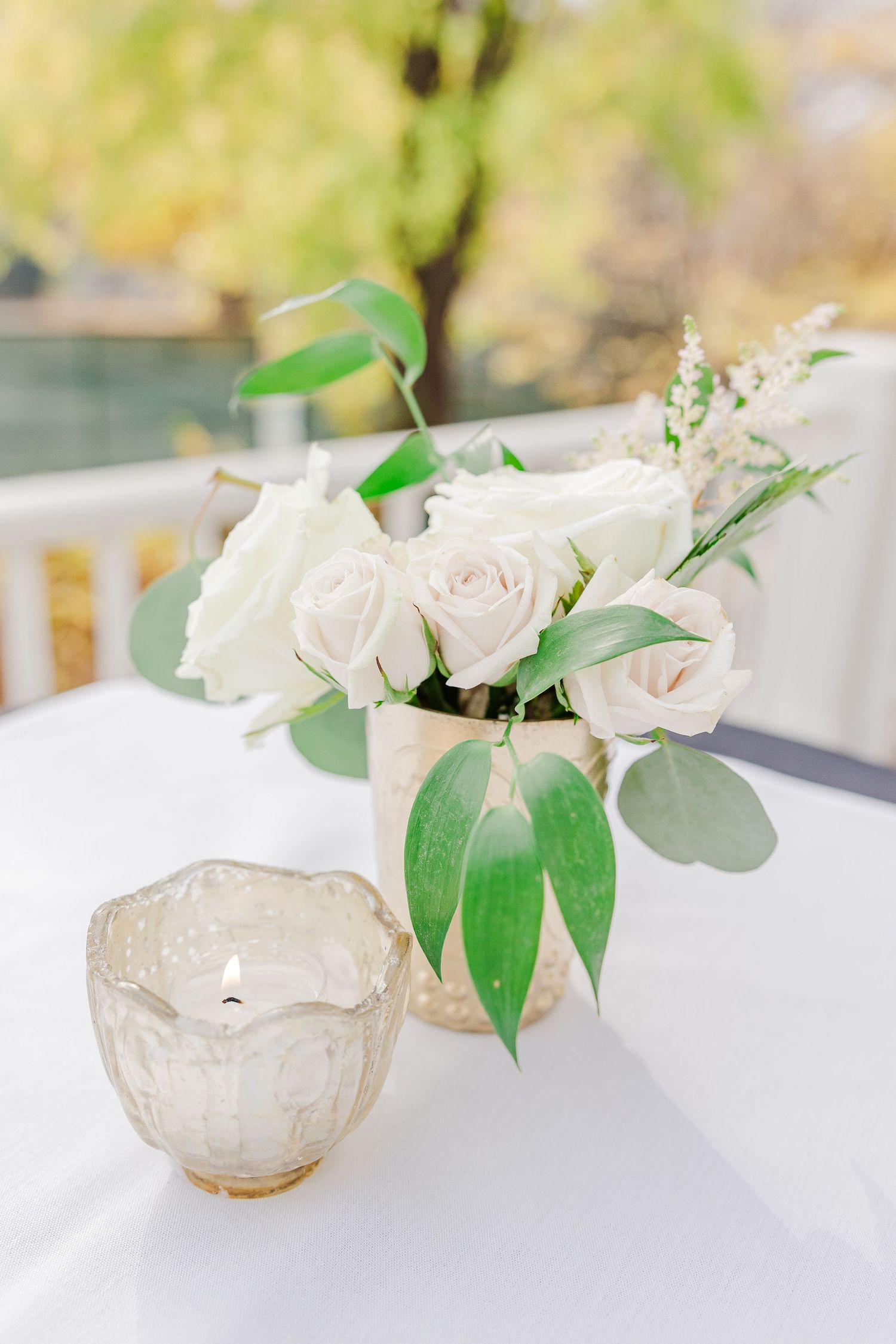 Elegant white rose centerpiece with eucalyptus in a glass vase next to a votice candle holder.