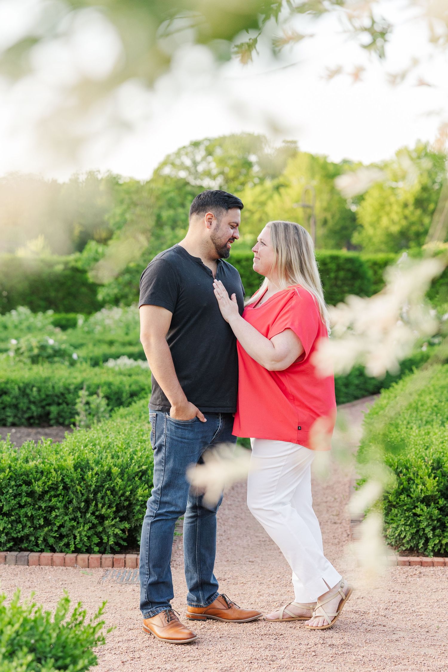 A couple shares a tender moment on a garden path surrounded by manicured hedges and greenery.