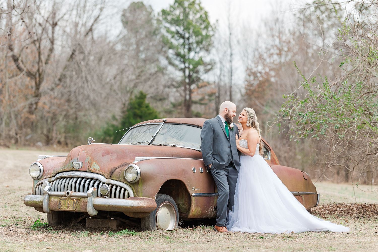 Newlyweds pose beside vintage rusty Buick in natural outdoor setting.