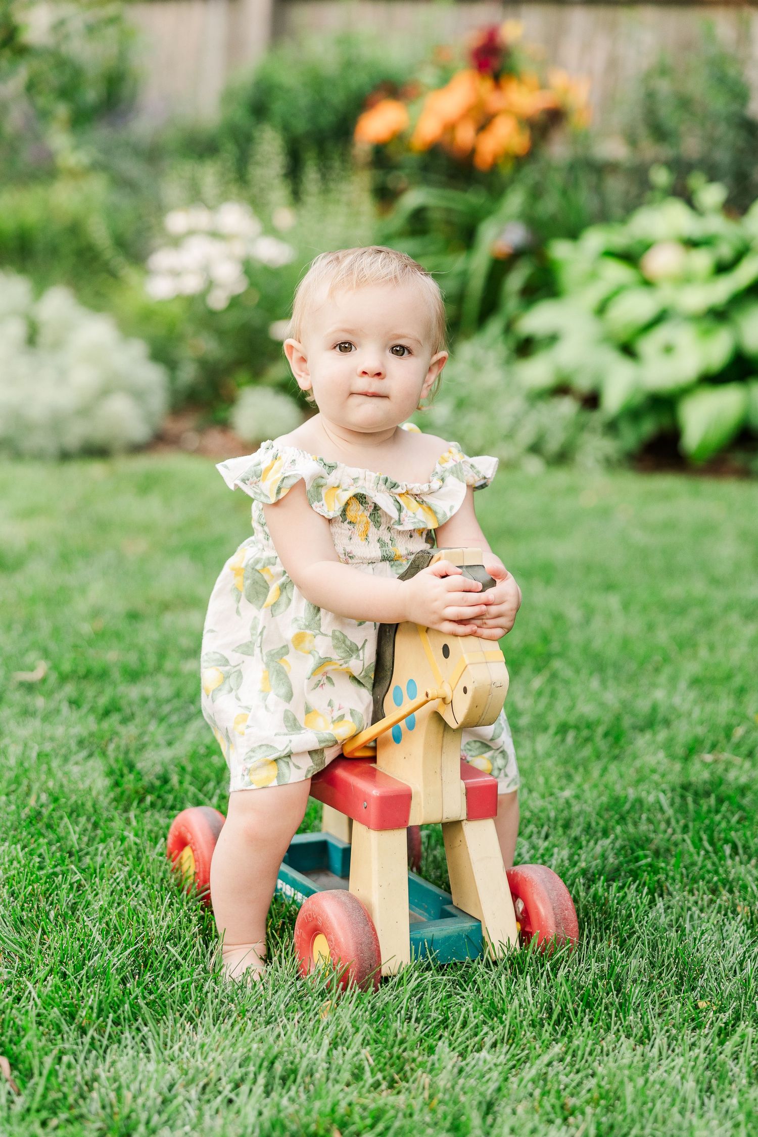 A young child sits on a colorful wooden ride-on toy surrounded by blooming garden flowers.