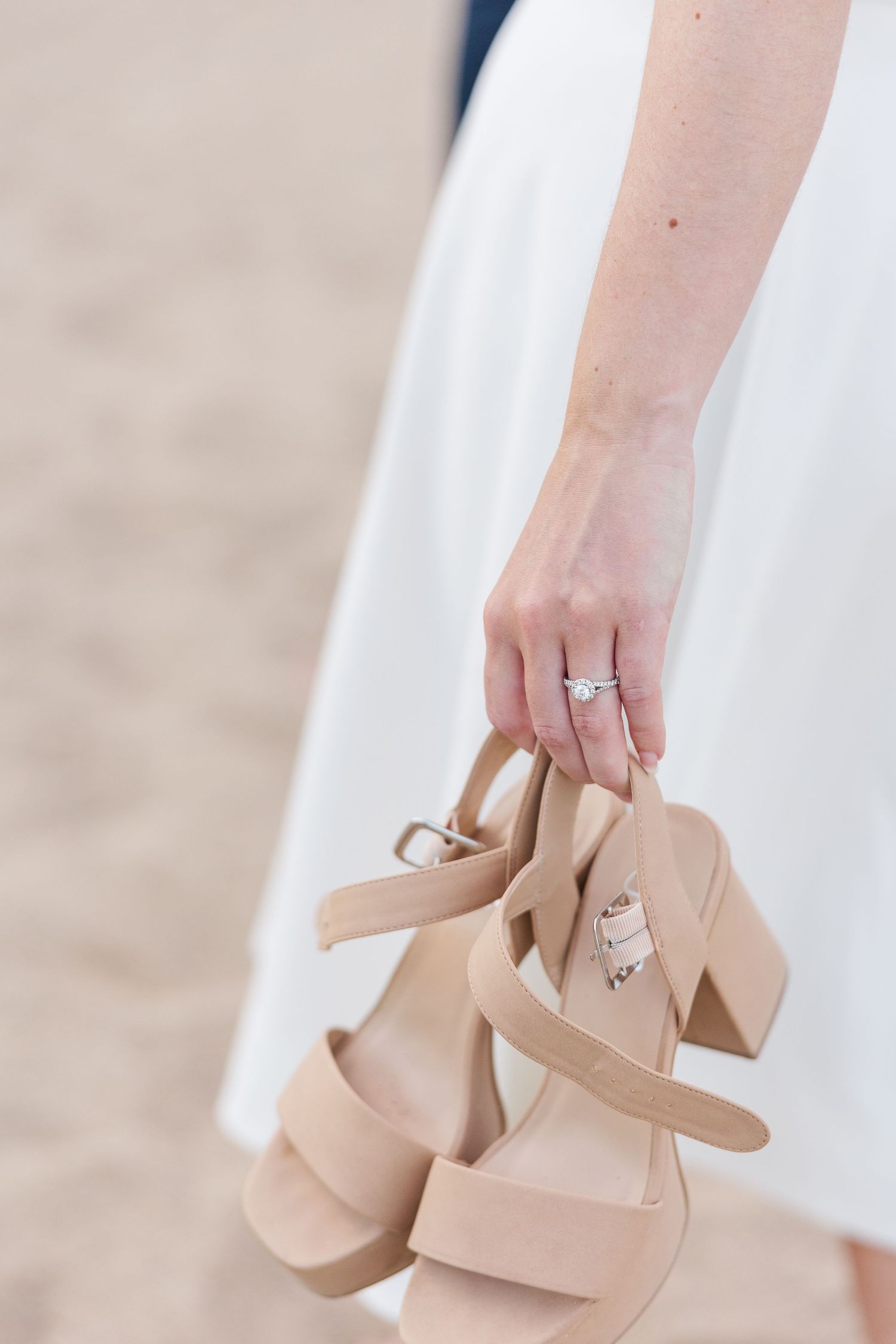 A hand holding nude-colored platform sandals while wearing a white dress and engagement ring.