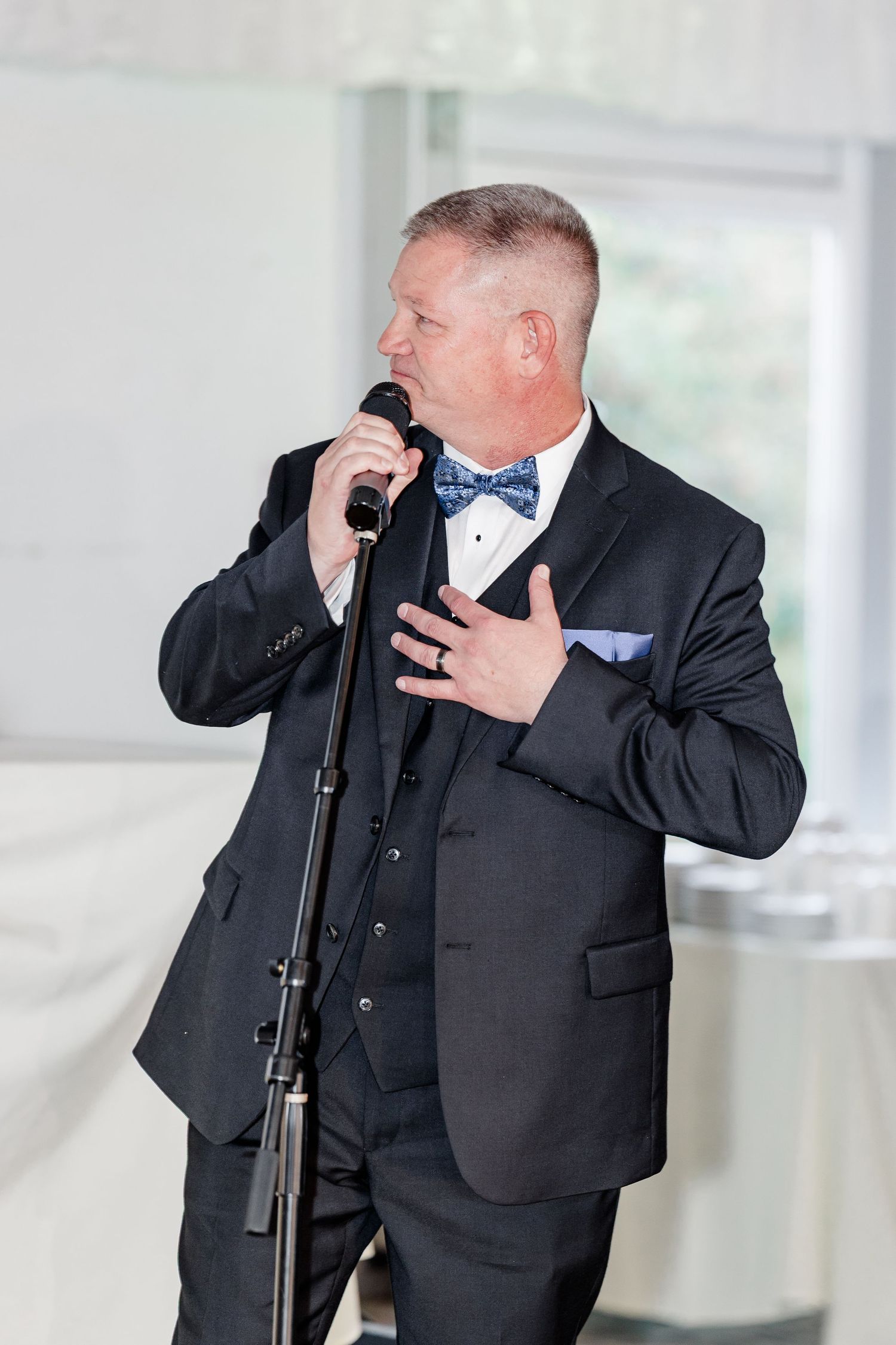 Man in black tuxedo with blue bow tie delivering a heartfelt speech at a wedding reception.