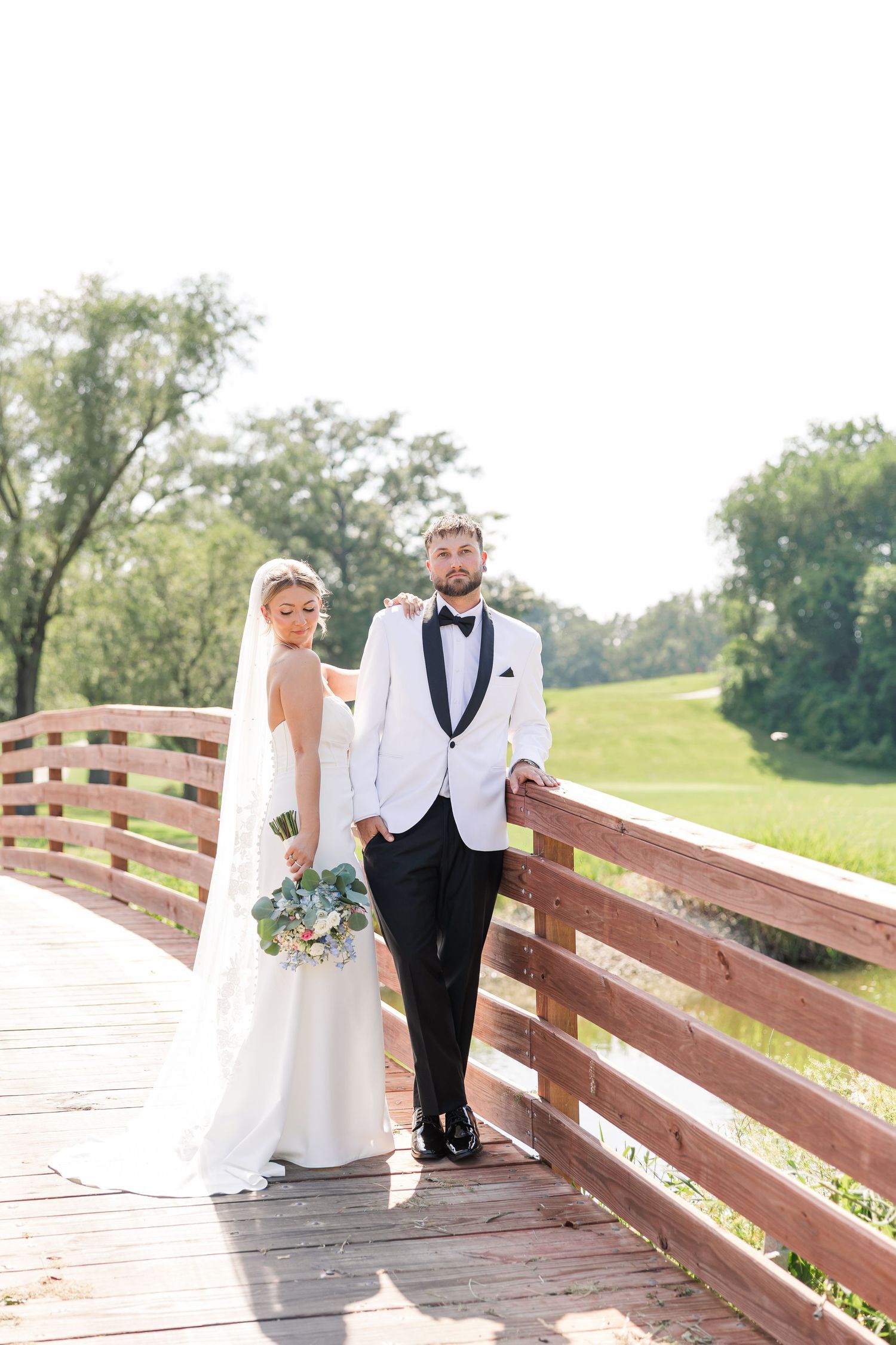 Bride and groom posing on rustic wooden bridge, groom in white tuxedo, bride holding bouquet.