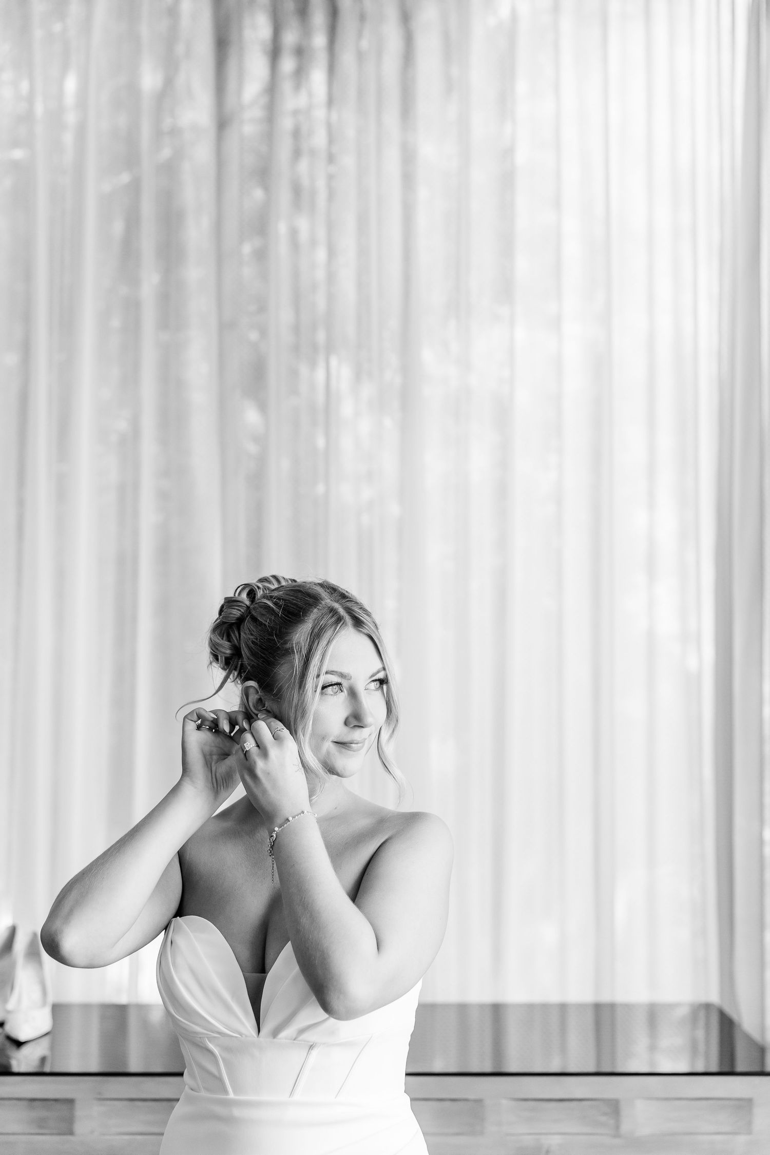 Black and white portrait of a bride in a strapless gown putting on earrings before her wedding.