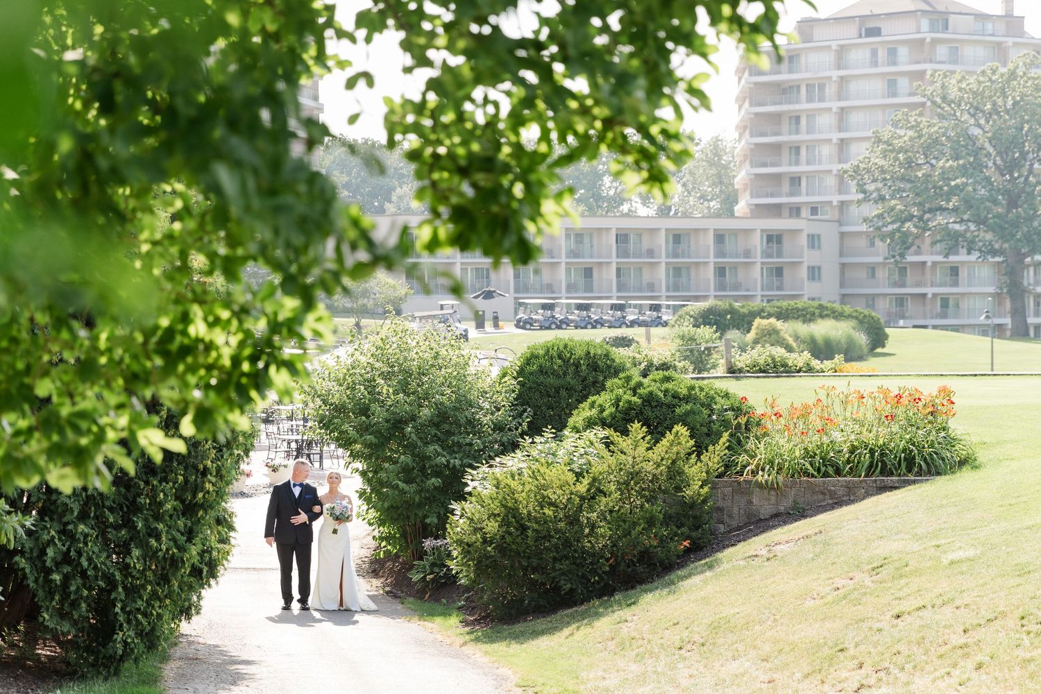 Bride and groom walking hand in hand through lush garden pathway with elegant hotel building in background.