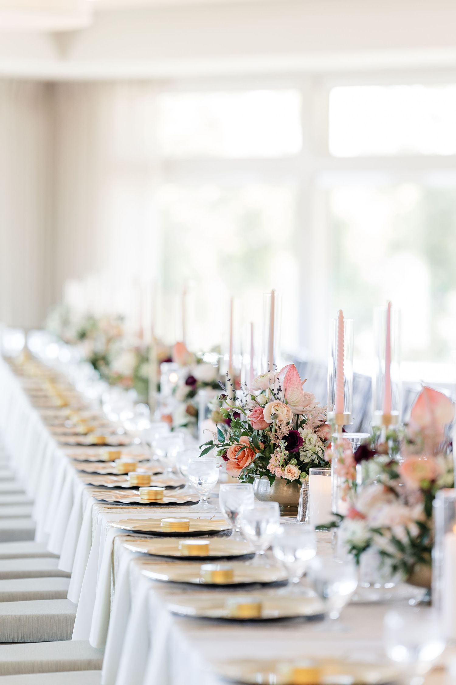 Elegant long wedding reception table with white linens, gold-rimmed plates, tall pink candles, and soft peach and blush floral centerpieces.