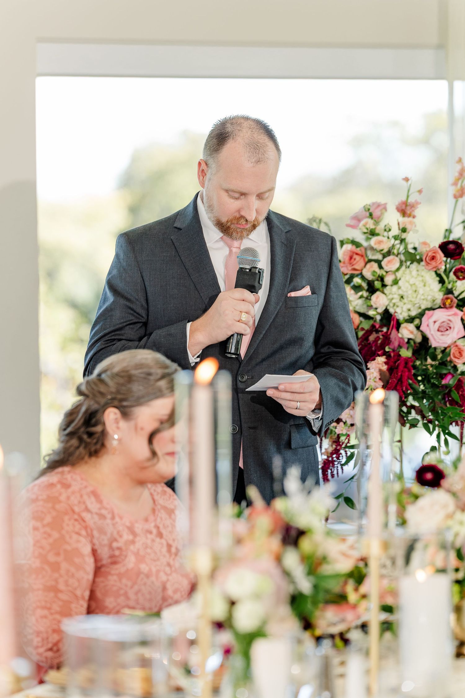 Man in a gray suit and pink tie reads a wedding speech into a microphone beside a colorful floral arrangement and candlelit table.
