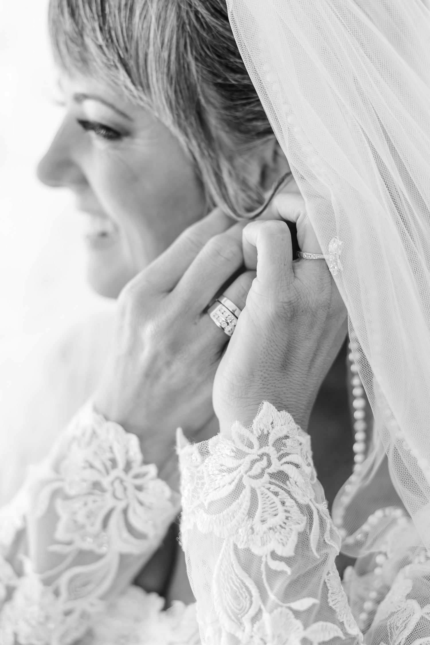 Black and white close-up of a smiling bride in a lace-sleeved wedding gown and veil adjusting her earring, wearing a ring.