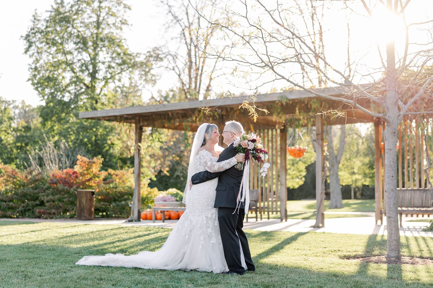 Bride in a flowing white gown and groom in a dark suit embrace holding a bouquet on a sunny lawn near a wooden pergola with autumn pumpkins.