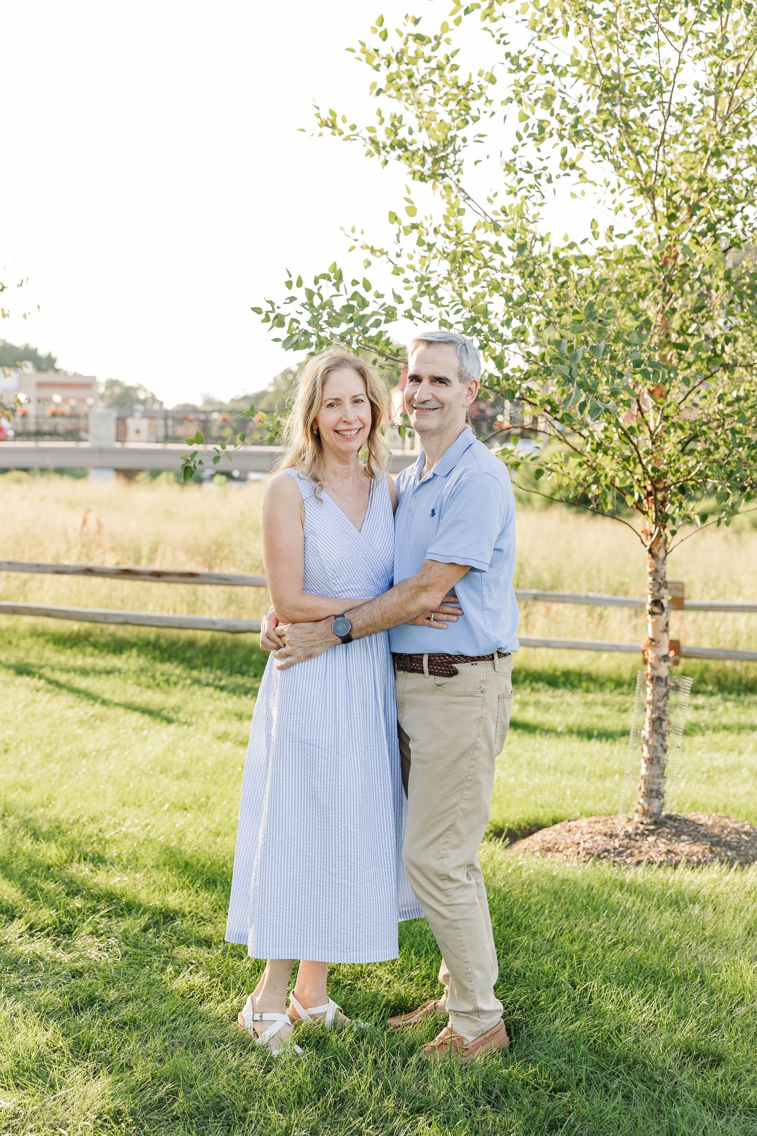 A smiling couple in casual attire embraces outdoors near a young tree on a sunny day.