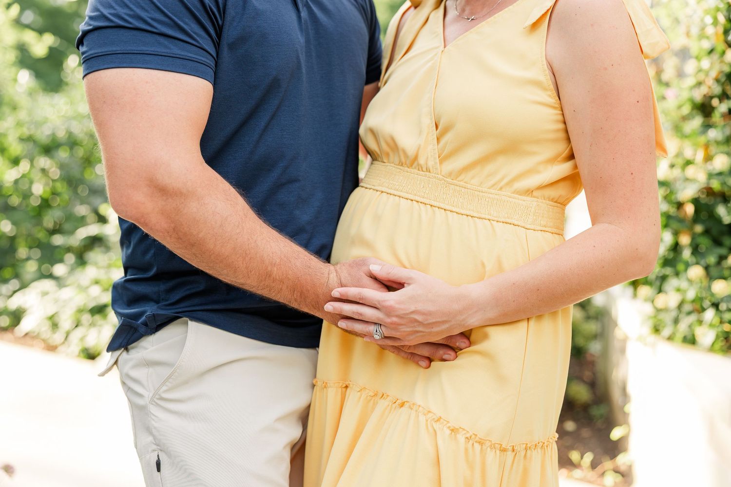 Close-up of couple cradling a pregnant belly outdoors, wearing navy and yellow outfits in garden.