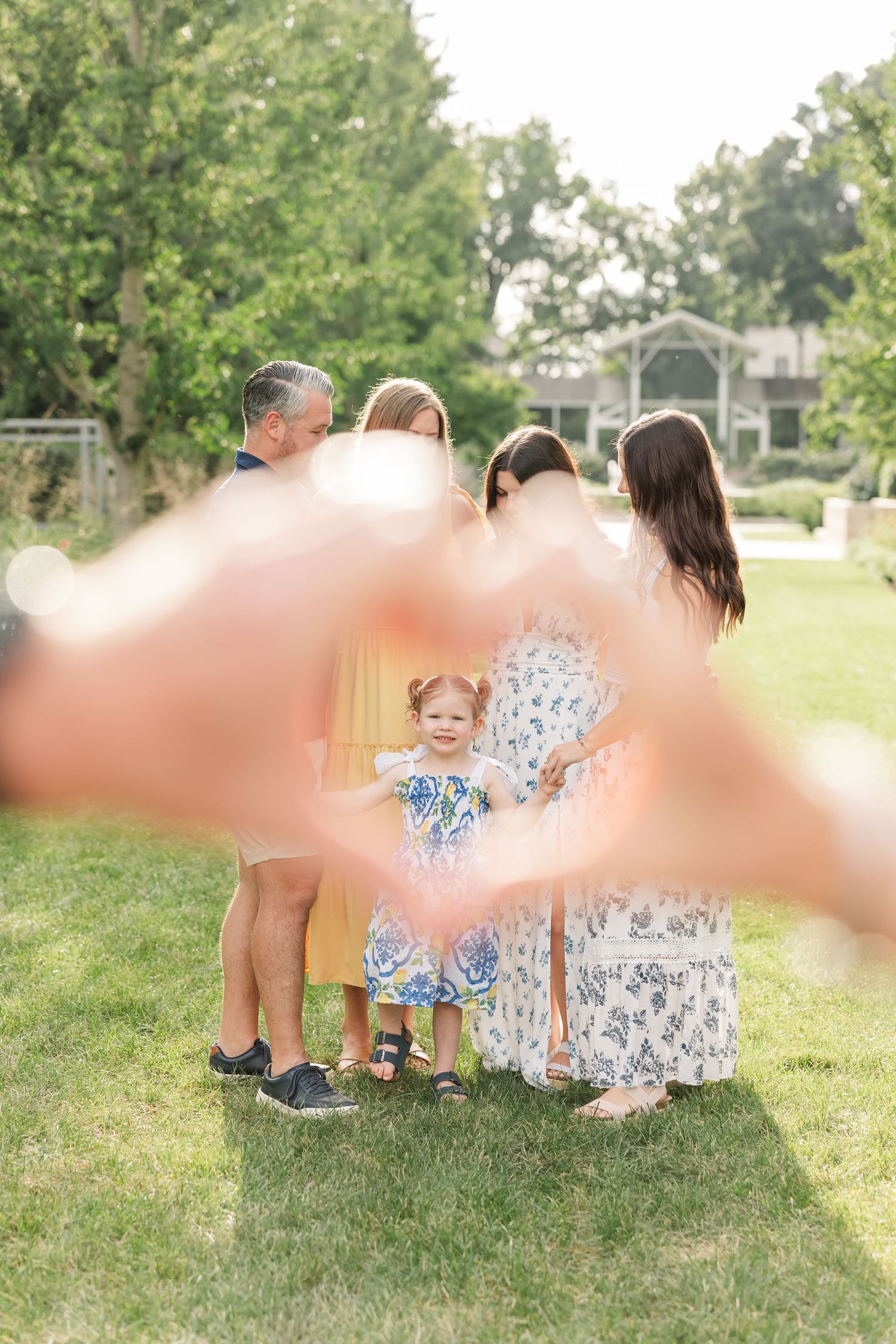Hands forming a heart frame around a family with a toddler girl on a green lawn.