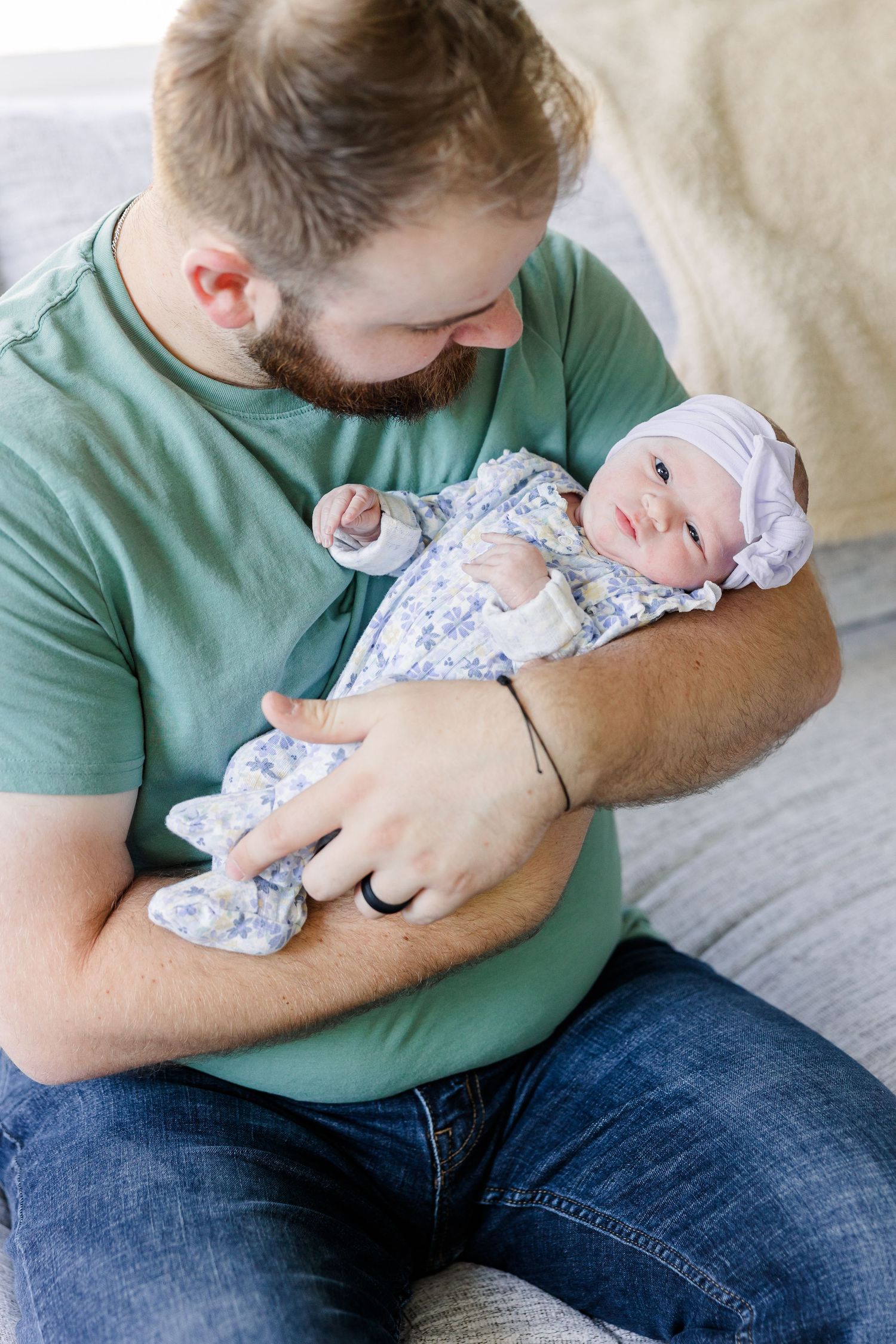 Bearded father in green shirt tenderly holds his newborn baby girl dressed in floral outfit with purple headband.
