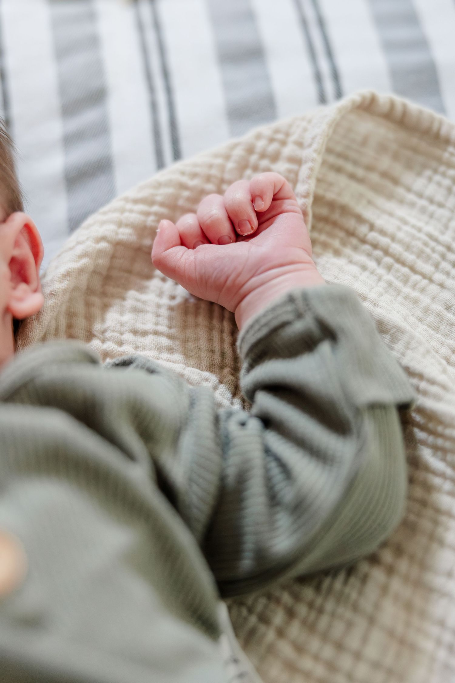 Close-up of a newborn baby's tiny fist resting on a cream textured blanket wearing a sage green outfit.