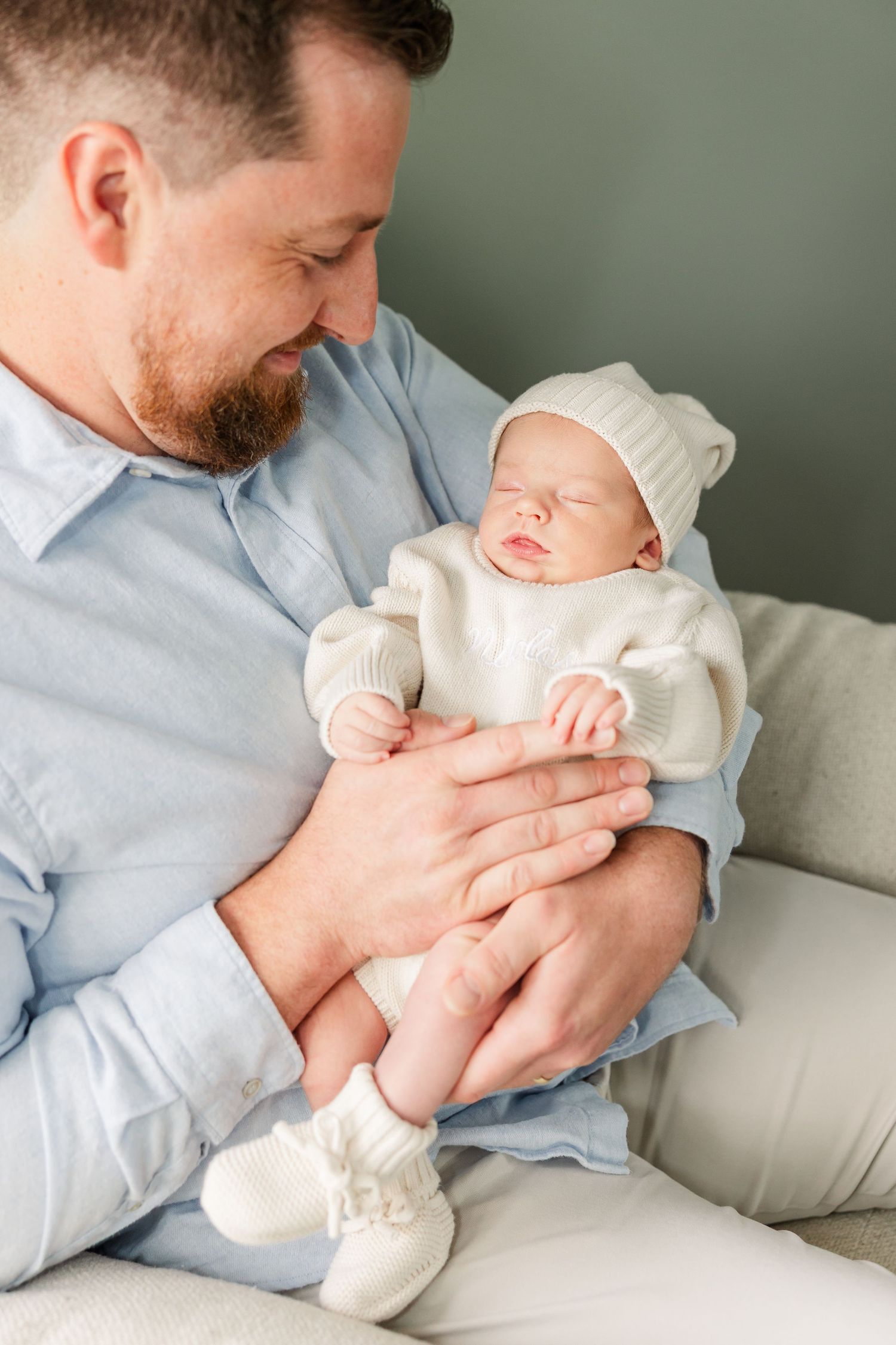 Bearded father in blue shirt tenderly holds sleeping newborn baby dressed in white knit outfit and hat.