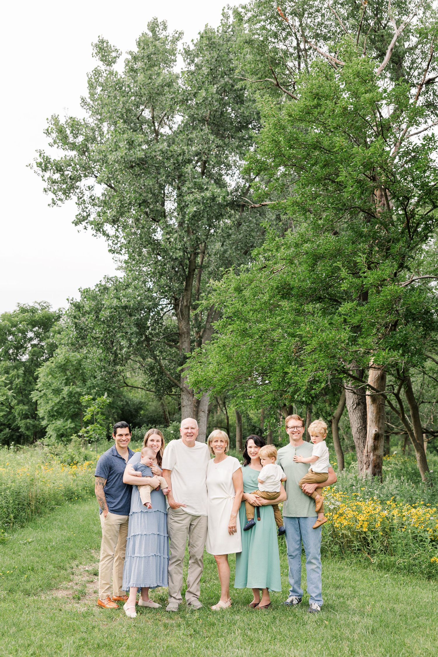 A multigenerational family of eight poses outdoors on a green lawn surrounded by lush trees wearing coordinated neutral tones.