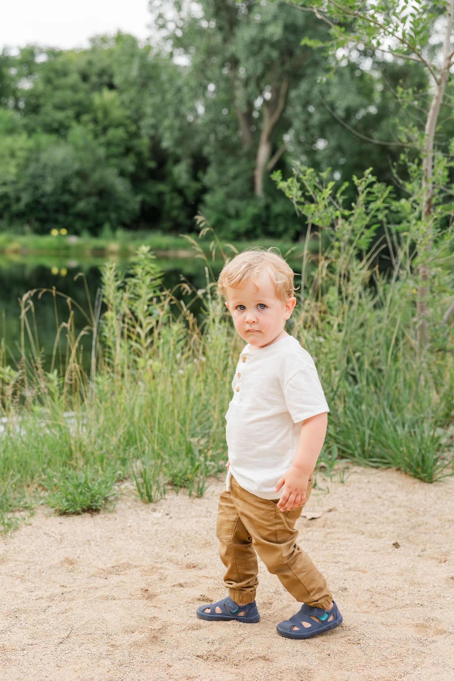 A blonde toddler boy in a white shirt and tan pants stands on sandy ground near lush green vegetation.
