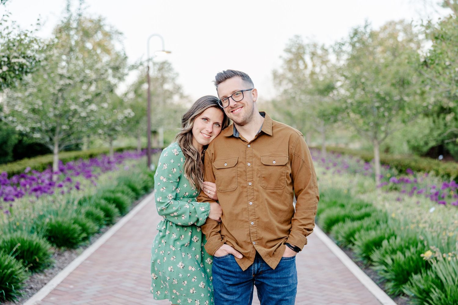 Smiling couple poses on a brick garden path surrounded by purple flowers and green trees.