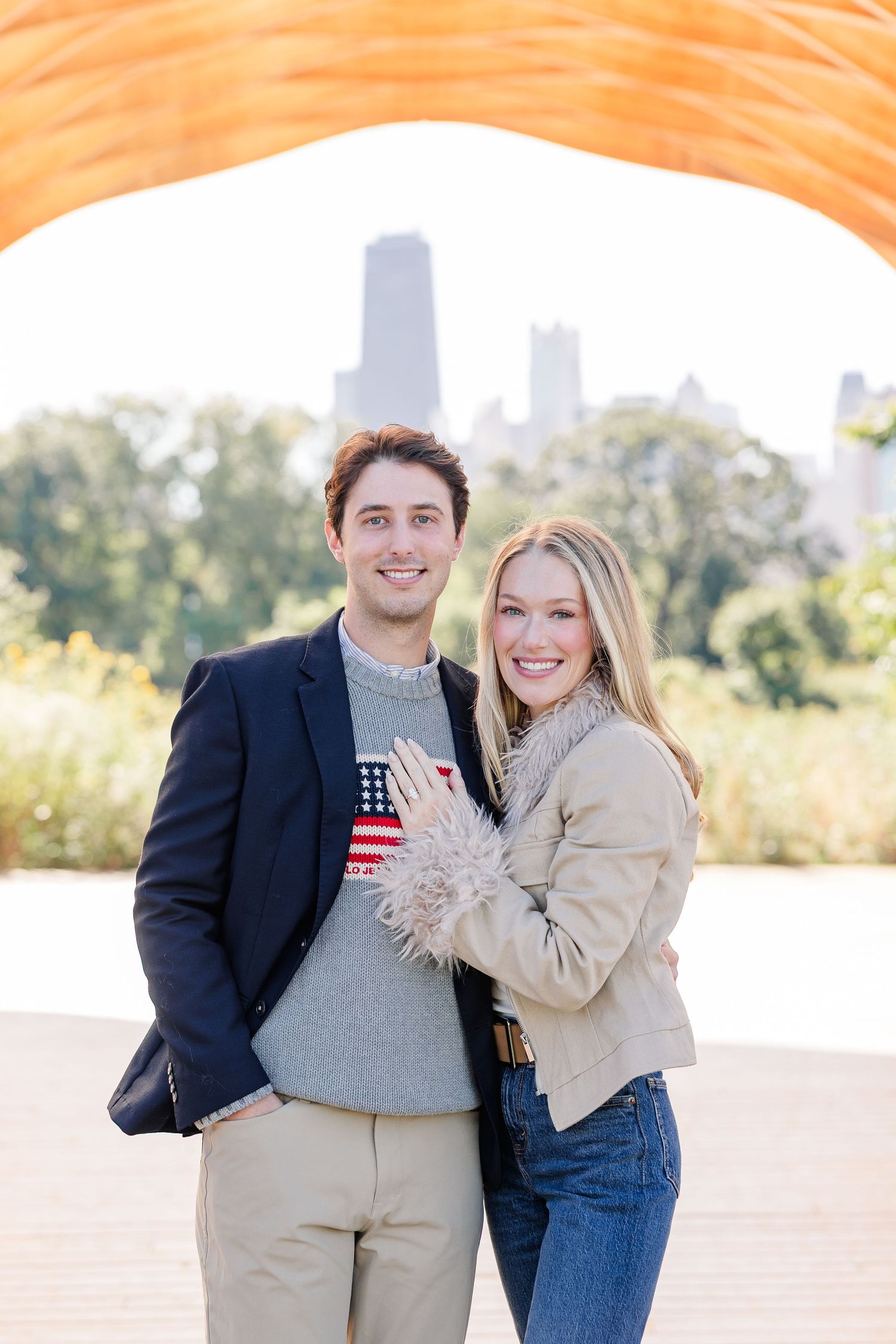Happy couple posing together under wooden arch structure with city skyline visible in background during engagement photos.