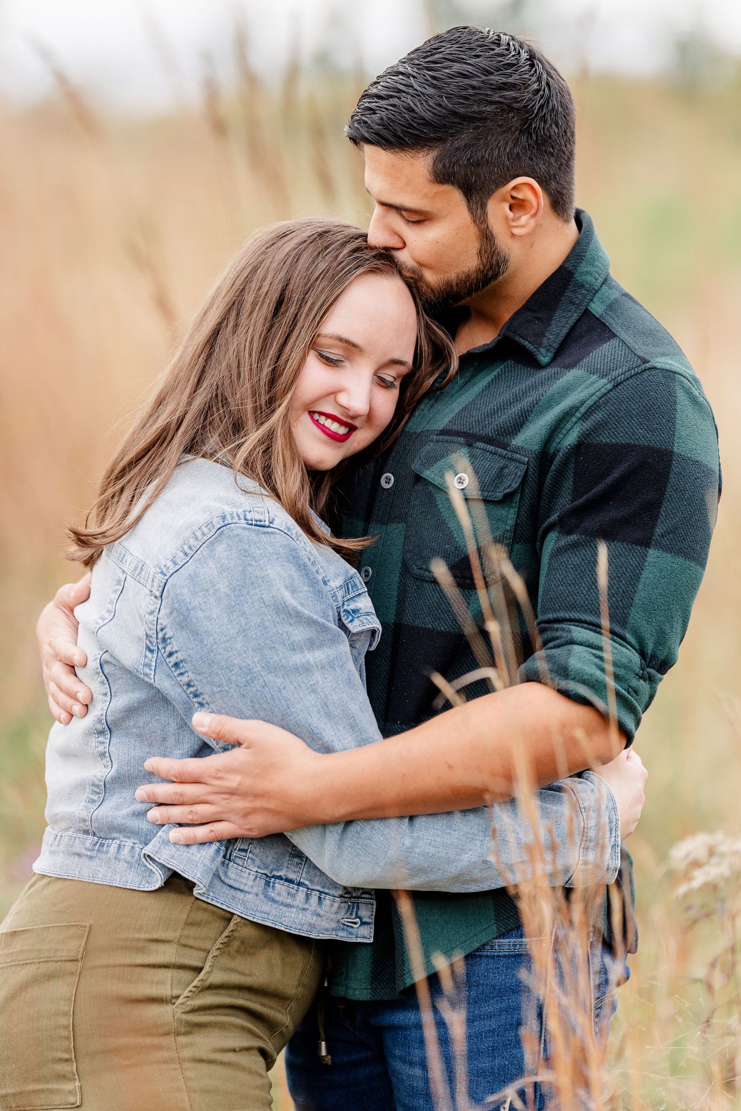 Man in green plaid shirt kisses a smiling woman's forehead as they embrace in a field of tall autumn grass.