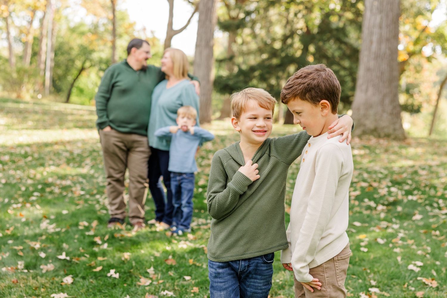 Two boys smile in a fall park while their family stands among autumn trees in the background.