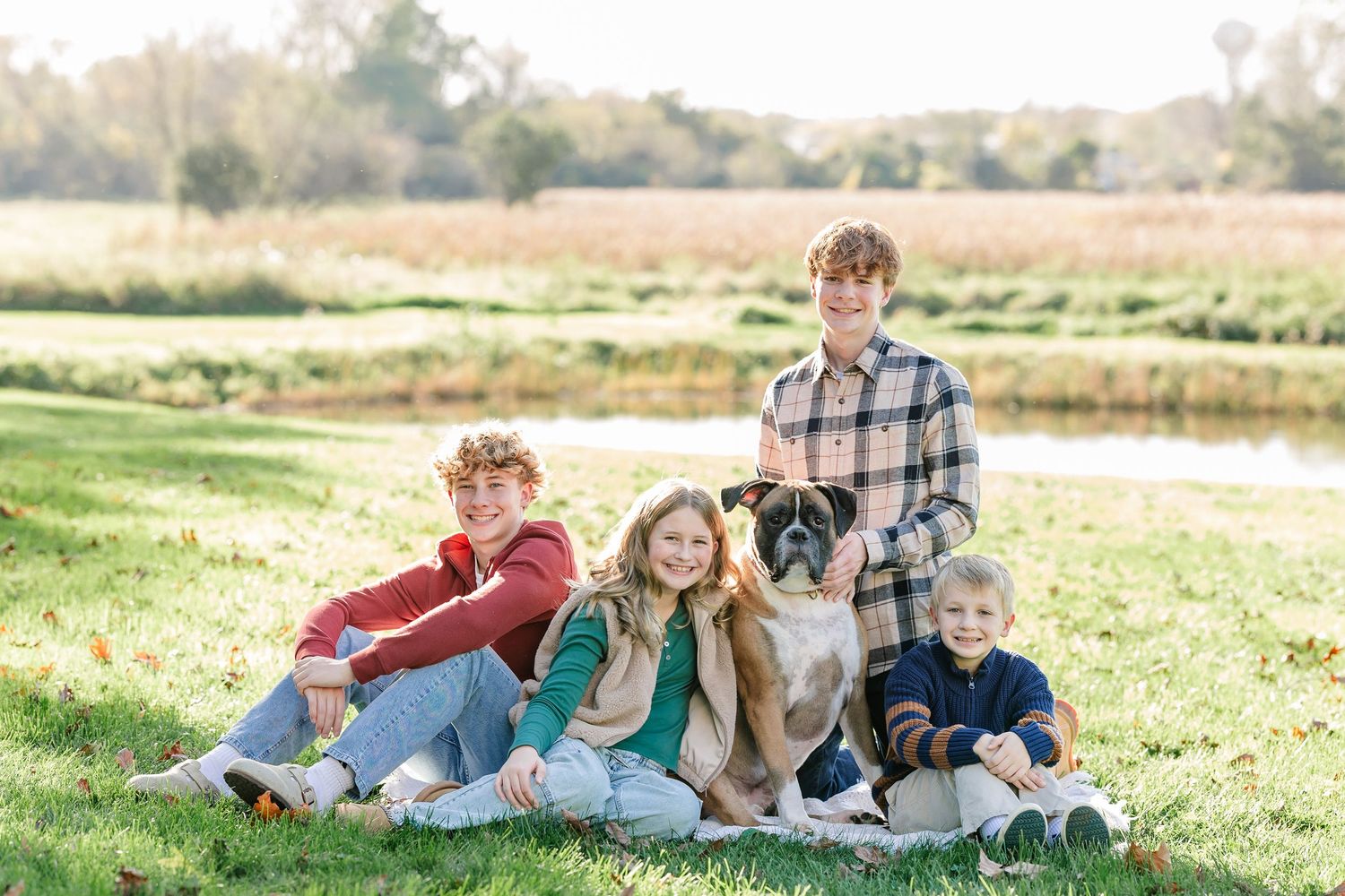 Four young family members sit and pose on a sunny grassy field with a boxer dog near a pond, wearing cozy fall clothing.