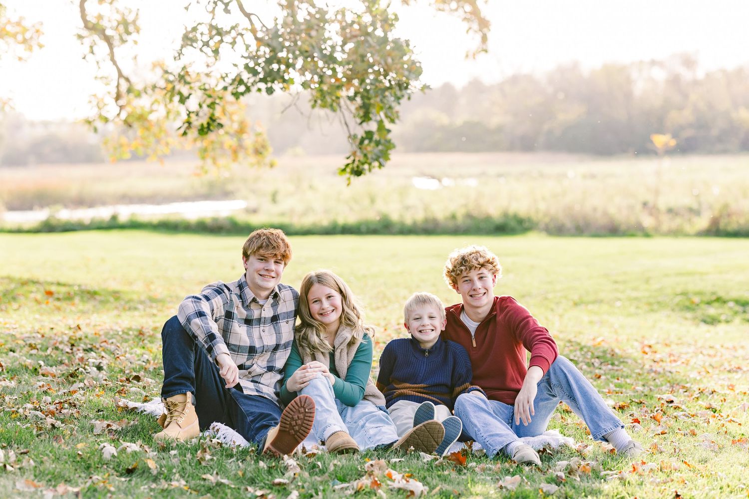Four young people sit close together on a grassy lawn dotted with fallen leaves, smiling under a tree with a soft autumn field behind them.