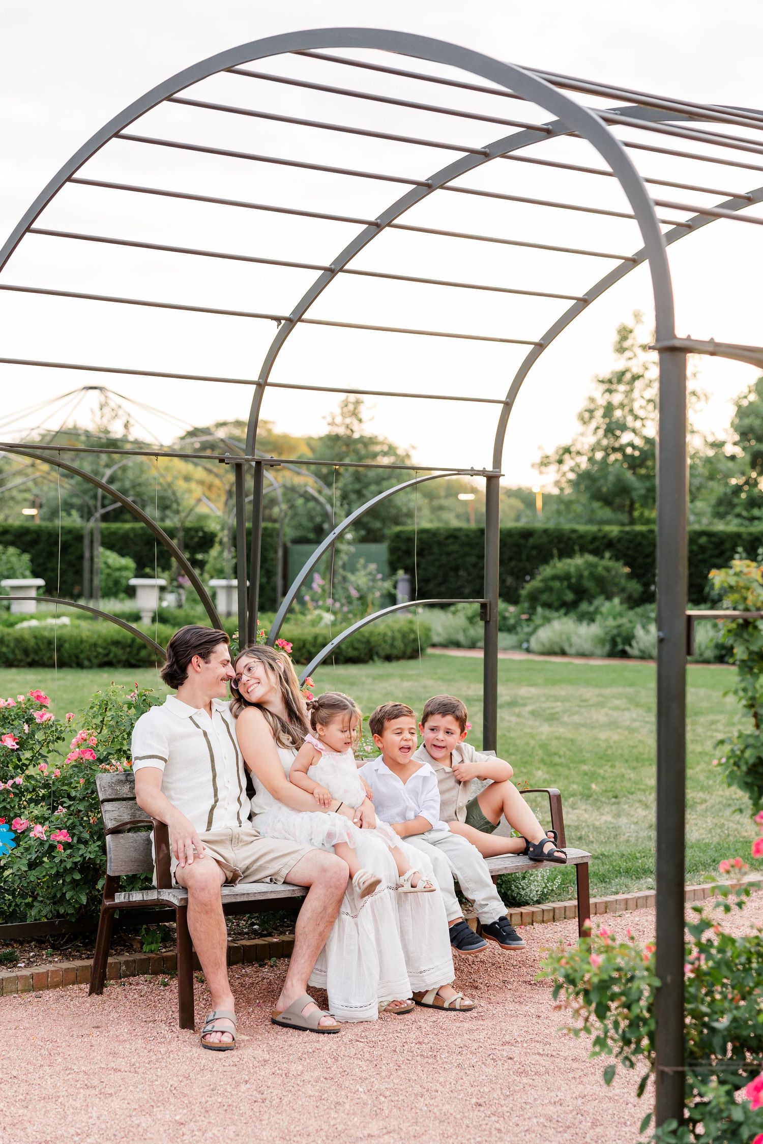 Happy family of five sits together on garden bench under metal arch surrounded by roses.