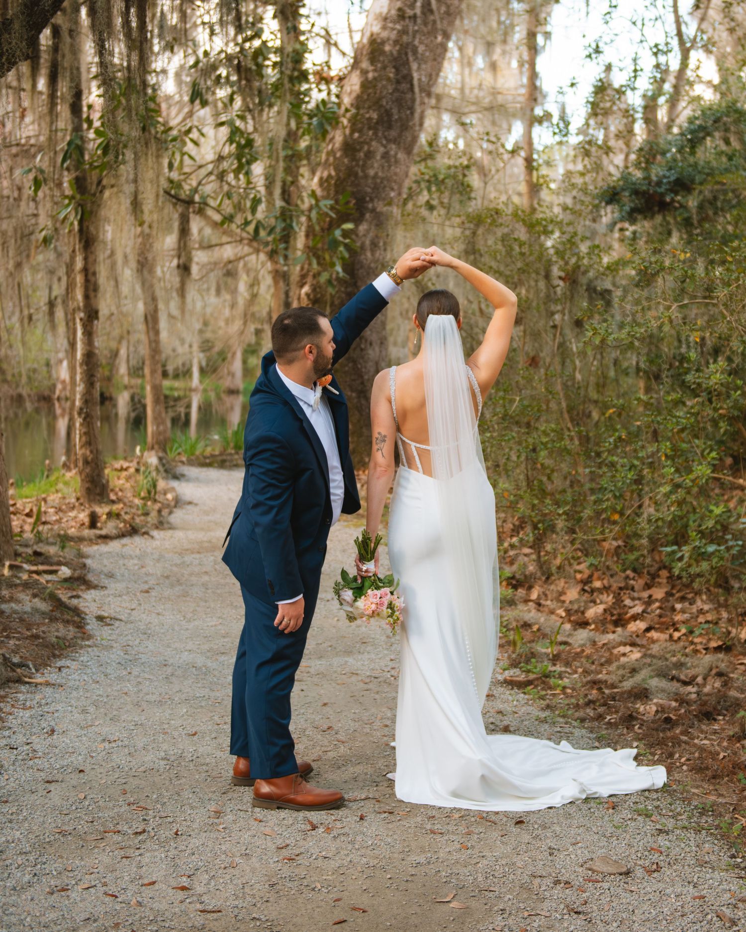 Bride twirling in her gown with groom beside her during a joyful, candid wedding moment under mossy trees.